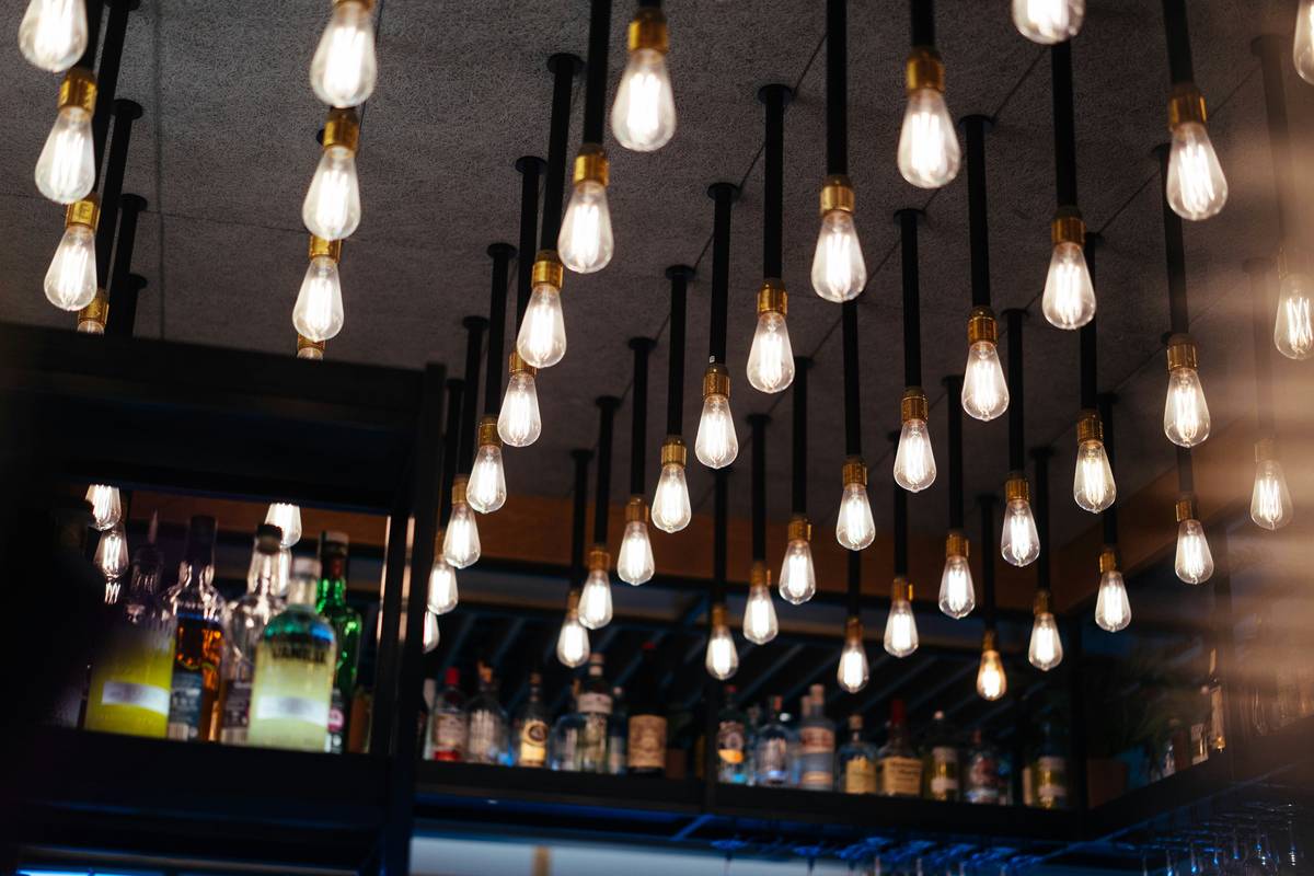 Side-by-side comparison: left shows harsh under-cabinet lighting casting shadows on bottles; right shows hanger bar fixture with even illumination highlighting liquid colors